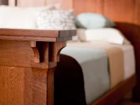 Detail shot of bedpost corner showing decorative wooden corbel bracket and layered crown molding in warm oak finish with soft bedding visible in background