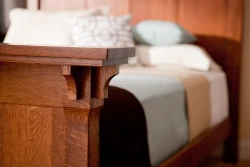 Detail shot of bedpost corner showing decorative wooden corbel bracket and layered crown molding in warm oak finish with soft bedding visible in background