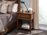 Transitional Shaker nightstand in brown maple featuring single drawer chrome pull open shelf tapered legs styled with industrial task lamp and white hydrangeas