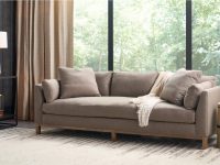 Rowe - Beige linen armchair with plush cushions, placed on a light rug near a wooden coffee table with a ceramic bowl and a potted plant by a large window