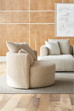 Rowe - Beige shaggy armchair with plush cushions, placed on a light rug alongside a beige sofa, with a floor lamp and framed artwork on a wooden paneled wall