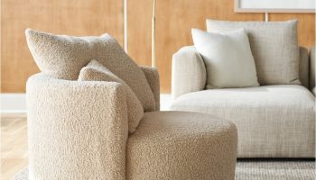 Rowe - Beige shaggy armchair with plush cushions, placed on a light rug alongside a beige sofa, with a floor lamp and framed artwork on a wooden paneled wall