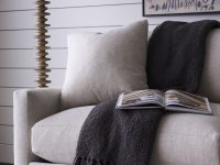 Rowe - Beige upholstered sofa with plush cushions and a dark gray throw, featuring an open book, placed near a tall wooden floor lamp and a framed artwork on a white shiplap wall