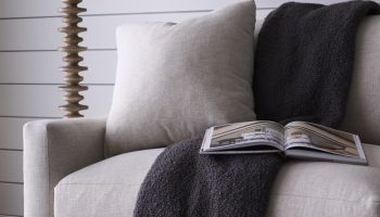 Rowe - Beige upholstered sofa with plush cushions and a dark gray throw, featuring an open book, placed near a tall wooden floor lamp and a framed artwork on a white shiplap wall