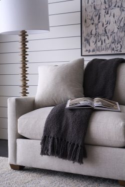 Rowe - Beige upholstered sofa with plush cushions and a dark gray throw, featuring an open book, placed near a tall wooden floor lamp and a framed artwork on a white shiplap wall