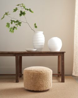 Rowe - Close-up view of a beige shaggy ottoman under a wooden console table, adorned with white textured vases and a branch with green leaves