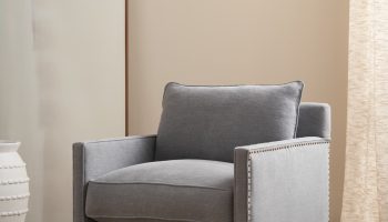 Rowe - Gray upholstered armchair with nailhead trim and wooden legs, placed on a beige rug near a large mirror and a white textured vase