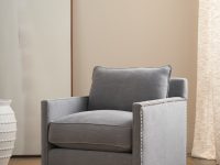 Rowe - Gray upholstered armchair with nailhead trim and wooden legs, placed on a beige rug near a large mirror and a white textured vase