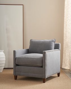 Rowe - Gray upholstered armchair with nailhead trim and wooden legs, placed on a beige rug near a large mirror and a white textured vase