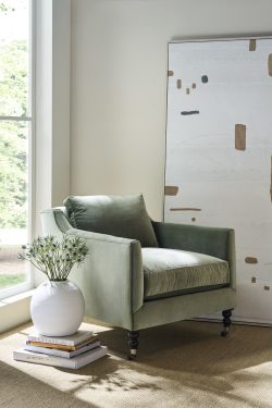 Rowe - Green velvet armchair with plush cushions, placed on a beige rug near a white vase with flowers and a stack of books by a large window