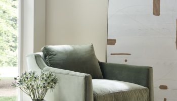 Rowe - Green velvet armchair with plush cushions, placed on a beige rug near a white vase with flowers and a stack of books by a large window