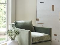 Rowe - Green velvet armchair with plush cushions, placed on a beige rug near a white vase with flowers and a stack of books by a large window