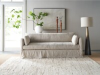 Rowe - close-up view of a beige linen sofa with plush cushions, placed on a light textured rug near a white vase with green branches, wooden sculptures, and a tall floor lamp
