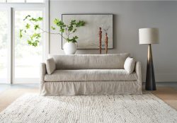 Rowe - close-up view of a beige linen sofa with plush cushions, placed on a light textured rug near a white vase with green branches, wooden sculptures, and a tall floor lamp