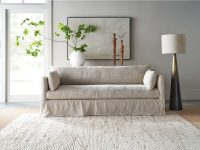Rowe - close-up view of a beige linen sofa with plush cushions, placed on a light textured rug near a white vase with green branches, wooden sculptures, and a tall floor lamp