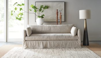 Rowe - close-up view of a beige linen sofa with plush cushions, placed on a light textured rug near a white vase with green branches, wooden sculptures, and a tall floor lamp