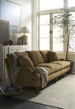 Rowe - close-up view of a tan upholstered sofa with plush cushions and a draped throw, placed on a light rug near a white cabinet with a lamp, books, and a potted olive tree by a large window