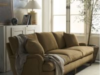 Rowe - close-up view of a tan upholstered sofa with plush cushions and a draped throw, placed on a light rug near a white cabinet with a lamp, books, and a potted olive tree by a large window