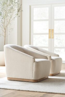 Rowe - close-up view of two beige upholstered armchairs with wooden bases, placed on a light rug near a potted olive tree and a large window with white doors