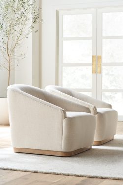 Rowe - close-up view of two beige upholstered armchairs with wooden bases, placed on a light rug near a potted olive tree and a large window with white doors