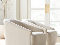 Rowe - close-up view of two beige upholstered armchairs with wooden bases, placed on a light rug near a potted olive tree and a large window with white doors