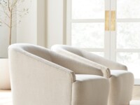 Rowe - close-up view of two beige upholstered armchairs with wooden bases, placed on a light rug near a potted olive tree and a large window with white doors