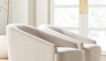 Rowe - close-up view of two beige upholstered armchairs with wooden bases, placed on a light rug near a potted olive tree and a large window with white doors